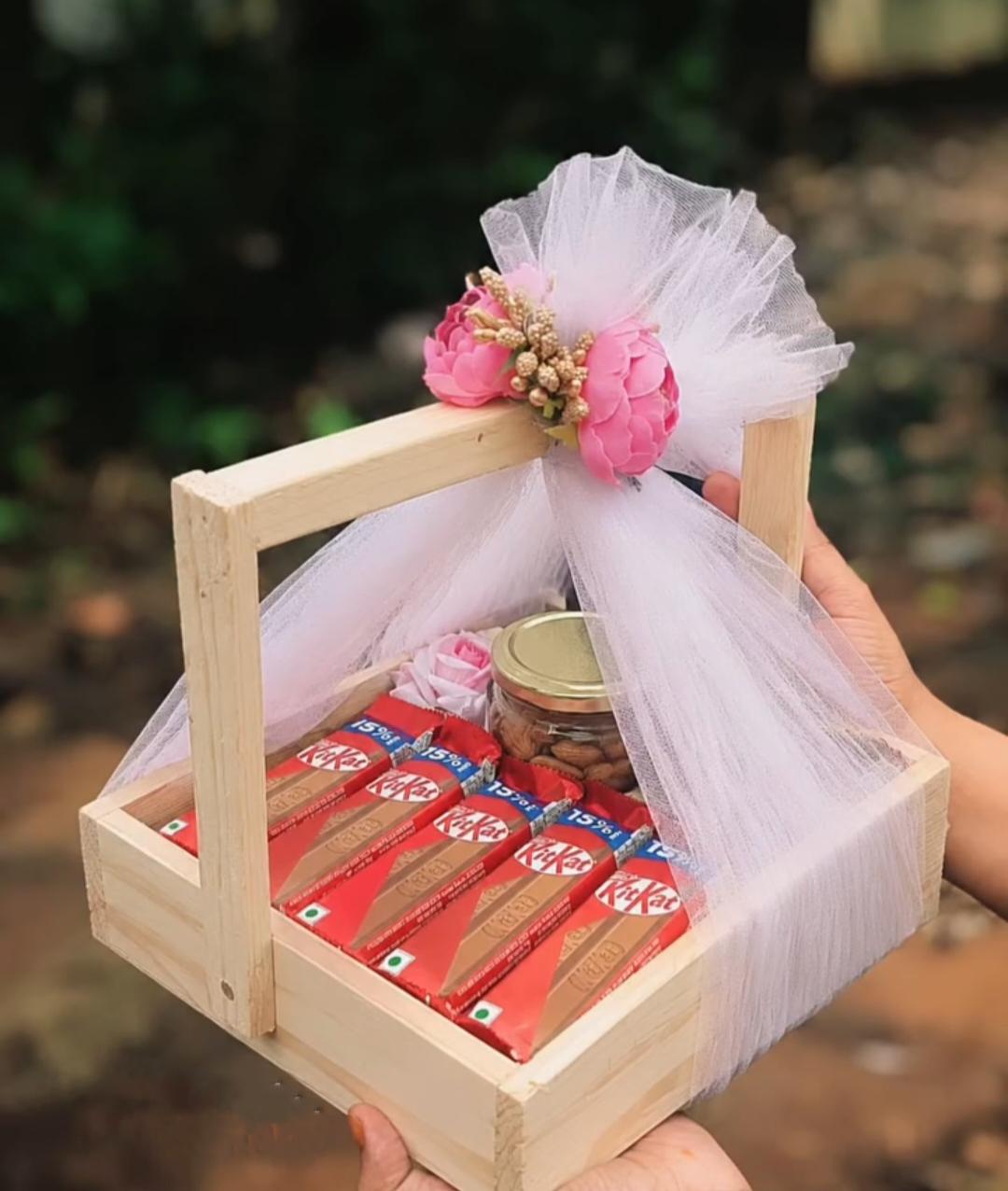 A wooden gift hamper with chocolates, a nut jar, pink flowers, and tulle